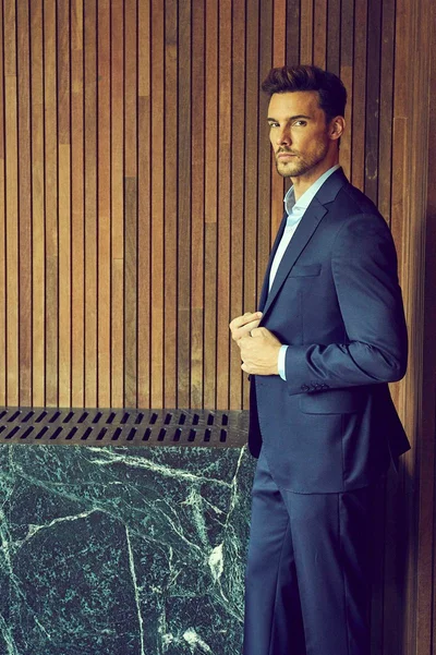 Man wearing a navy suit jacket and trousers posing against a vertical wooden slat wall and dark green marble counter.