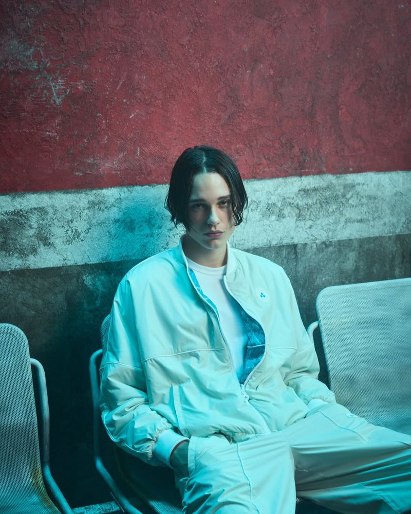 Portrait of a young adult wearing a white tracksuit sitting in an indoor setting with a distressed red and gray wall in the background.