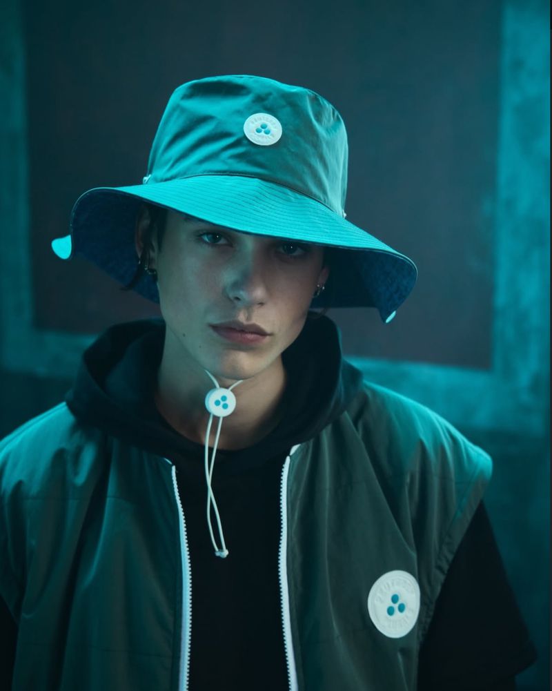Portrait of a young adult wearing a blue bucket hat and olive green vest in a studio setting with a dark teal background.