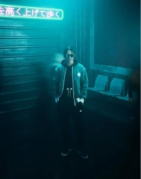 Model wearing a teal jacket and dark clothing under a glowing neon sign in a moody urban alleyway at night.