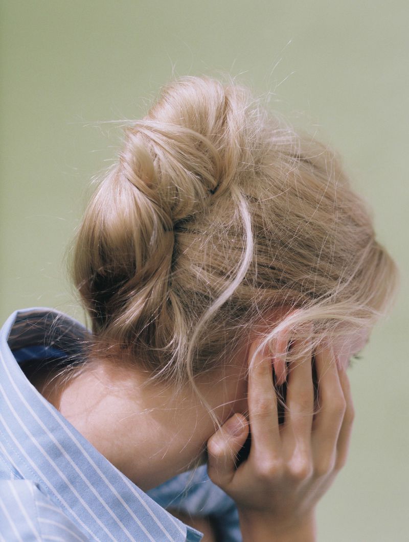 Close-up on a blonde woman with a bun, wearing a blue striped shirt, with her face obscured by her hand against a pale green background.