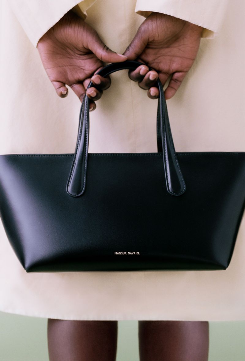 Black leather tote bag held by hands. Minimalist fashion accessory detail, studio product shot for online retail, beauty brand, or ad campaign.