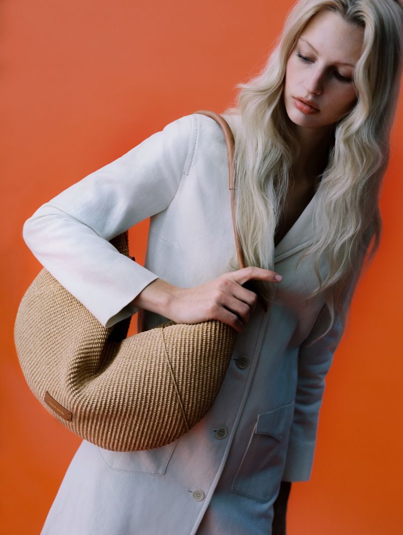 A blonde woman poses in front of an orange background while showcasing a tan woven crescent-shaped purse with a light-colored strap.