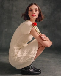 Model wearing cream knit dress and black boots crouching while holding red flower against textured grey backdrop