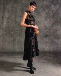 Model with short dark hair wearing a sleeveless sheer black dress and lace-up boots holding flowers against a grey backdrop.