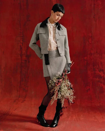 Model standing against a red textured backdrop wearing a grey tweed jacket, matching skirt, and holding dried flowers.