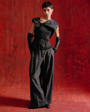 Model Krini Hernandez in a structured black top, wide-leg grey trousers, and long black gloves against a red textured backdrop.