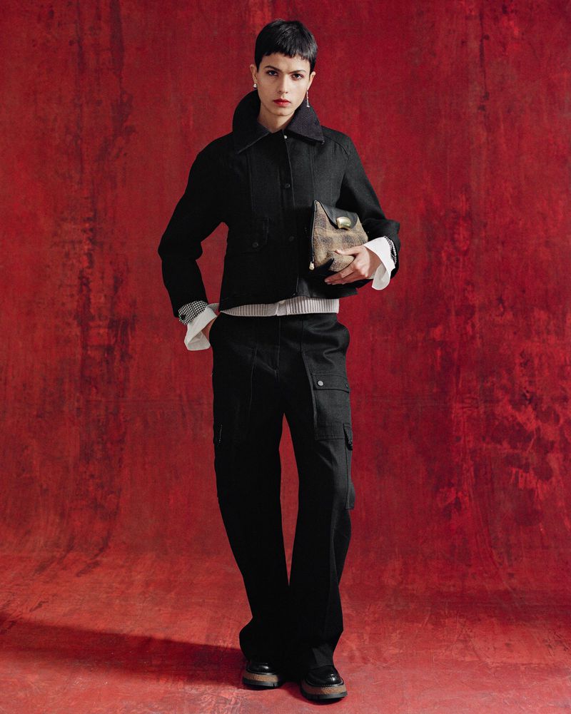 A fashionably dressed person in a dark jacket, light striped shirt, and cargo pants poses against a textured dark red backdrop indoors.
