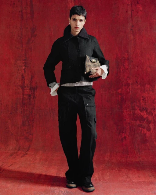 A fashionably dressed person in a dark jacket, light striped shirt, and cargo pants poses against a textured dark red backdrop indoors.