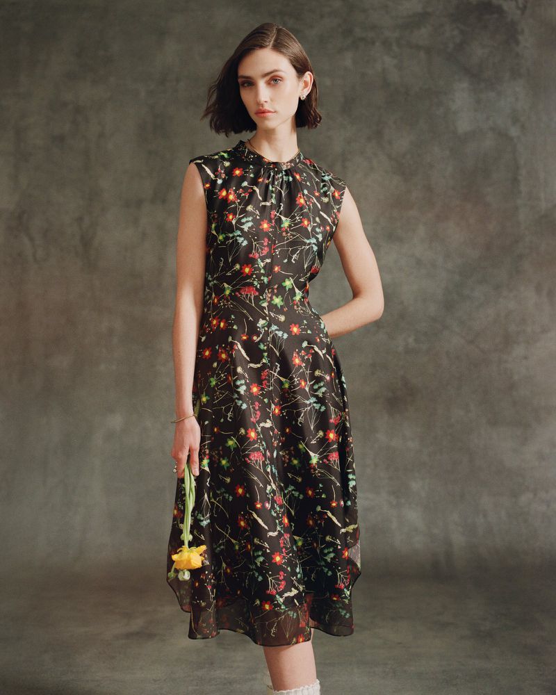 Woman in floral dress holds a yellow flower against a grey mottled backdrop in a studio with diffused lighting, fashion photography.