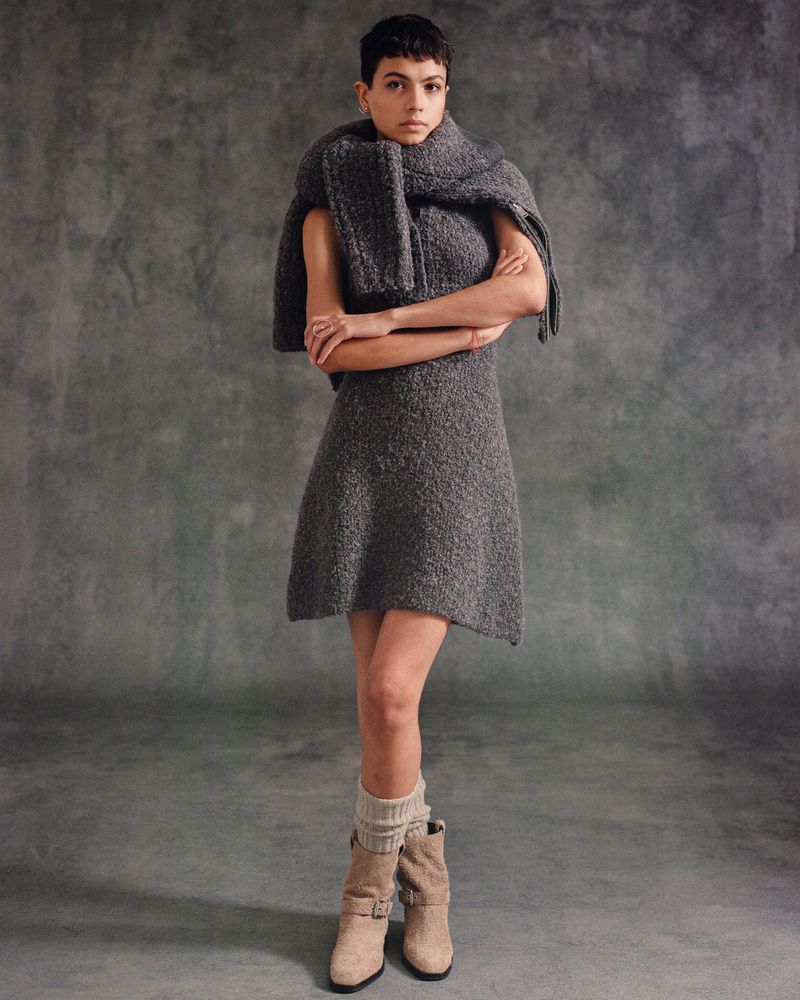 Woman in gray textured mini dress with draped scarf and cuffed boots poses in a studio with a mottled gray backdrop.