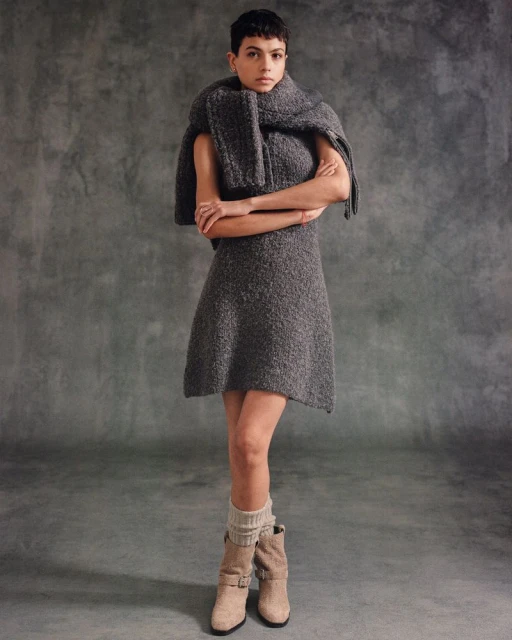 Woman in gray textured mini dress with draped scarf and cuffed boots poses in a studio with a mottled gray backdrop.