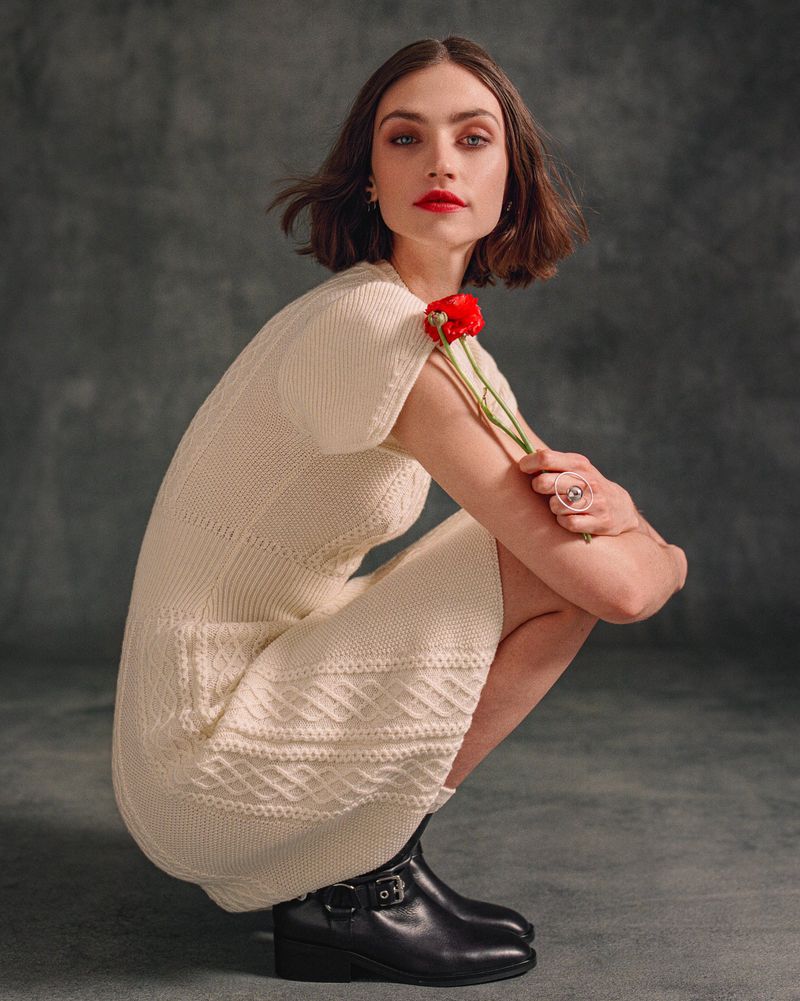 Portrait of a young woman in a cream cable-knit dress crouching with a red flower, set against a dark grey textured backdrop.