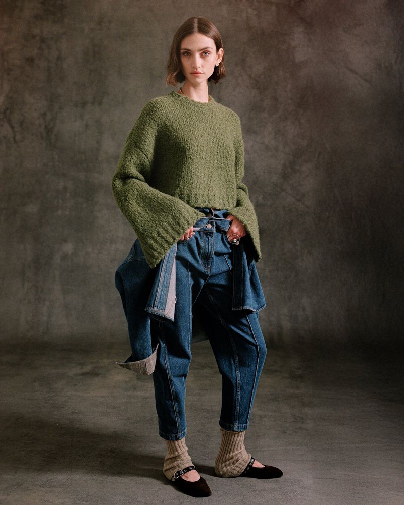 Full-length shot of a female fashion model in a green sweater, blue jeans, black flats with leg warmers, standing in a photo studio.