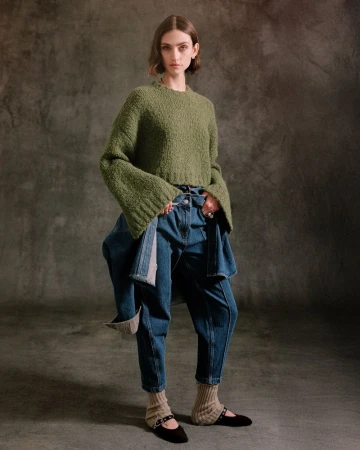 Full-length shot of a female fashion model in a green sweater, blue jeans, black flats with leg warmers, standing in a photo studio.