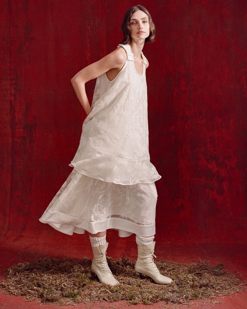 An adult female fashion model in a floor-length, white dress stands against a red backdrop with brown ground cover, illuminated by studio lighting.