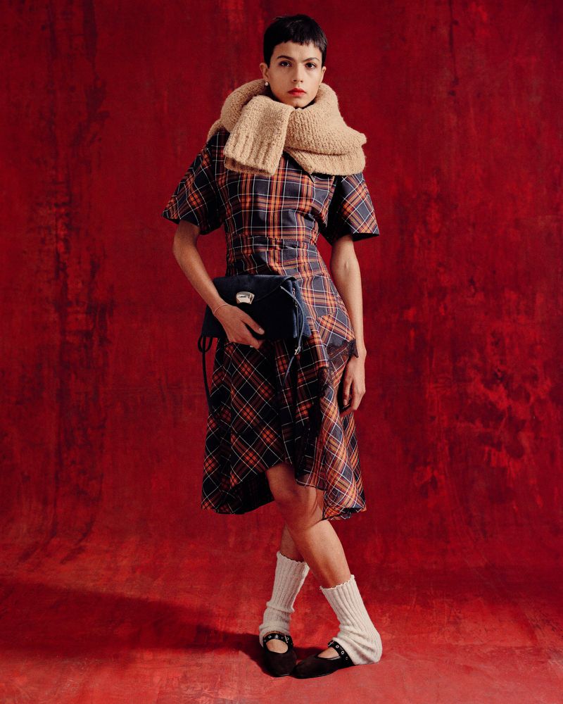 Fashion shot of a woman in a plaid dress, chunky knit scarf, Mary Jane shoes, and knee-high socks against a textured red background.