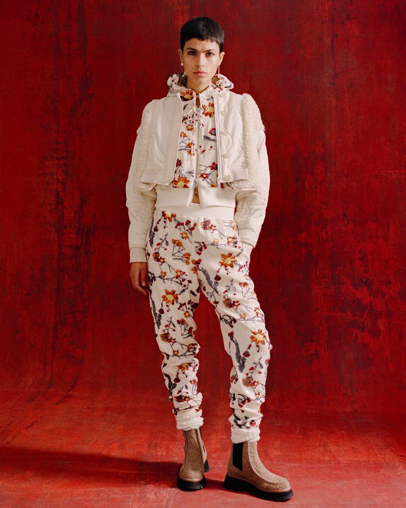 A woman in a patterned outfit poses against a red backdrop, showcasing a cropped jacket, floral top, and matching pants with boots.