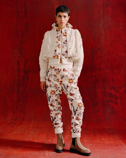 A woman in a patterned outfit poses against a red backdrop, showcasing a cropped jacket, floral top, and matching pants with boots.