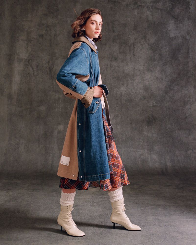 A woman in a patchwork denim and tan trench coat poses in front of a grey backdrop wearing off-white boots and a red plaid skirt.