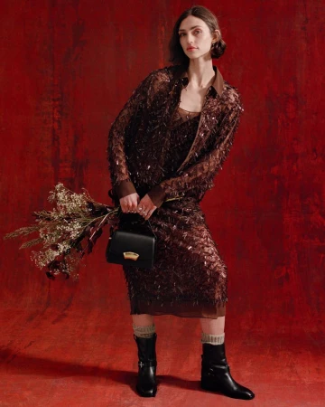 A stylish woman in a brown shimmering skirt suit holds flowers and a handbag against a textured red backdrop in a studio setting.
