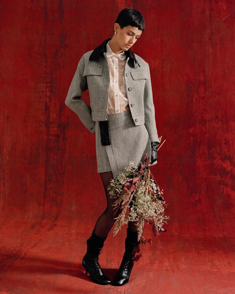A fashionably dressed woman poses against a red backdrop wearing a gray skirt suit, black boots, and sheer stockings while holding flowers.