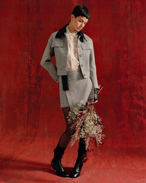 A fashionably dressed woman poses against a red backdrop wearing a gray skirt suit, black boots, and sheer stockings while holding flowers.