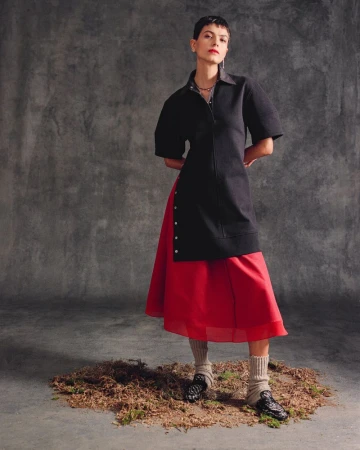 Model posing in a black short-sleeved long tunic layered over a vibrant red skirt against a textured grey backdrop.