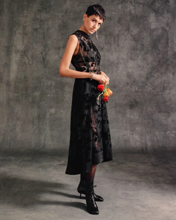 Model with short dark hair wearing a sleeveless sheer black dress and lace-up boots holding flowers against a grey backdrop.
