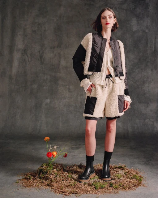 Woman models a quilted jacket and fleece shorts with ankle boots in a studio setting. She stands on moss near flowers.