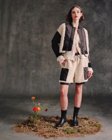 Model wearing a black and cream shearling jacket with matching fleece shorts, standing on a patch of moss against a grey backdrop.