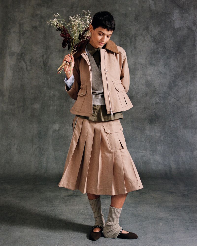 A woman in a tan jacket and skirt holding flowers poses in front of a mottled gray background, looking off to the side.