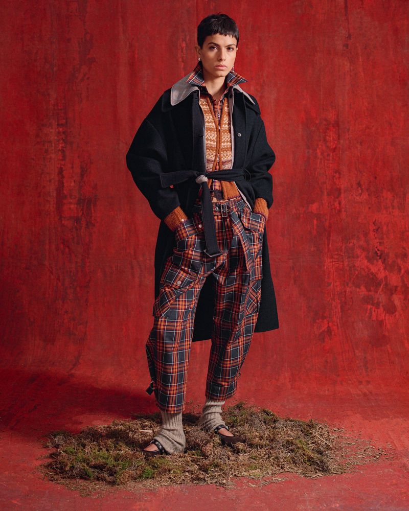 A woman poses in a patterned shirt and trousers with a black coat against a red background, standing on a mossy surface.