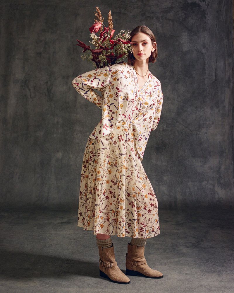 A stylish woman poses indoors with a bouquet, wearing a floral print dress, socks, and boots against a textured grey backdrop.