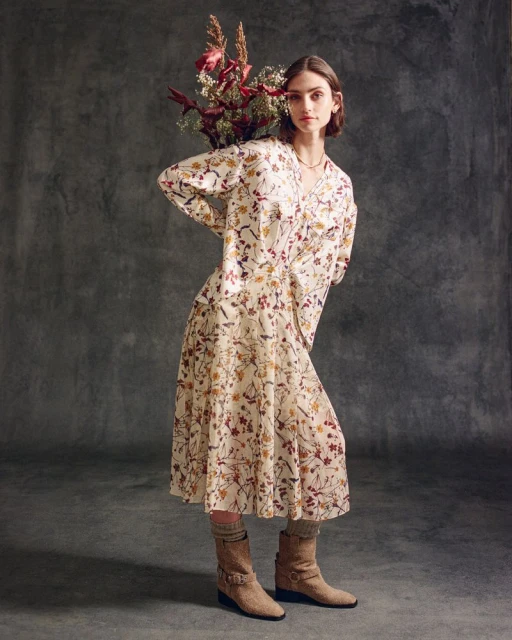 A stylish woman poses indoors with a bouquet, wearing a floral print dress, socks, and boots against a textured grey backdrop.
