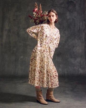 A stylish woman poses indoors with a bouquet, wearing a floral print dress, socks, and boots against a textured grey backdrop.