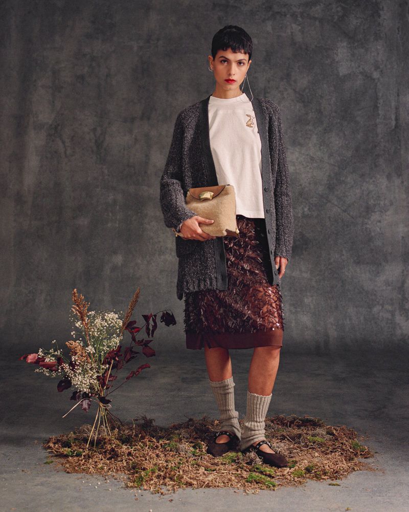 A stylish young woman poses in a studio wearing a gray cardigan, a short brown skirt, and a white t-shirt, holding a tan handbag.
