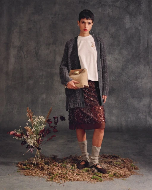 A stylish young woman poses in a studio wearing a gray cardigan, a short brown skirt, and a white t-shirt, holding a tan handbag.