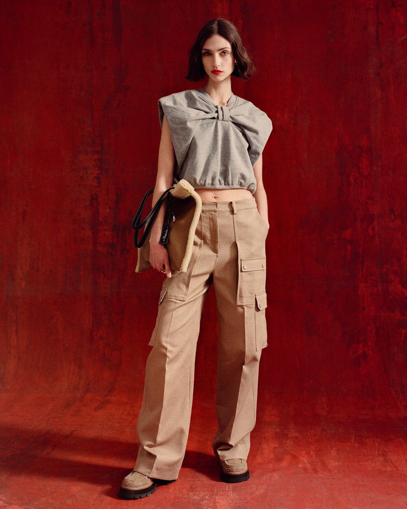 Fashion shot of a woman wearing a gray sleeveless top and khaki cargo pants, holding a bag, against a textured red background.