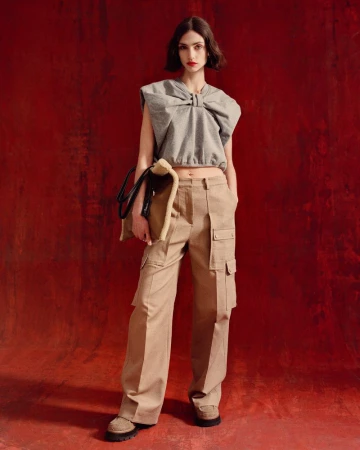 Fashion shot of a woman wearing a gray sleeveless top and khaki cargo pants, holding a bag, against a textured red background.