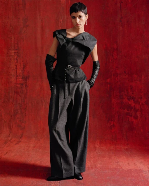 A female fashion model poses in a sleeveless dark top, wide grey pants, and black gloves against a textured red studio backdrop.