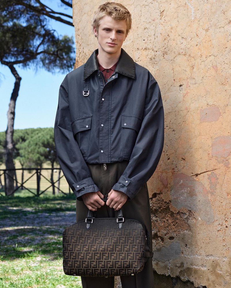 Male fashion model holds a Fendi handbag outdoors. He wears a blue-gray jacket, standing in front of a weathered wall and green foliage.