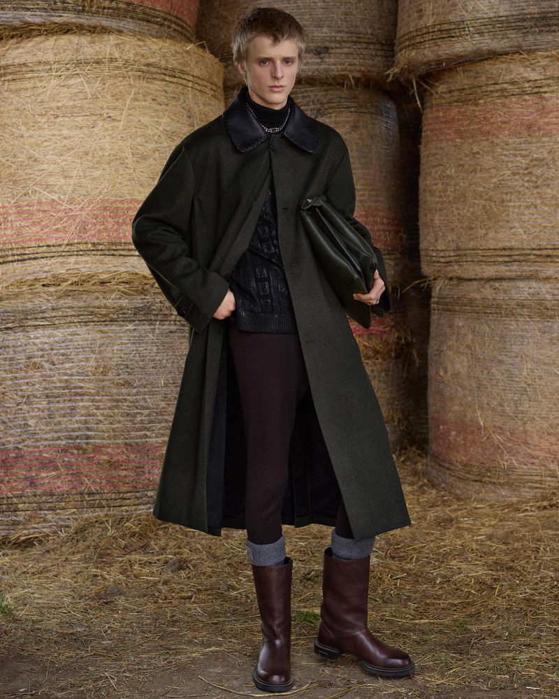 A fair-skinned adult male poses in a dark green coat, brown trousers, and boots, holding a dark green bag against a hay bale backdrop.