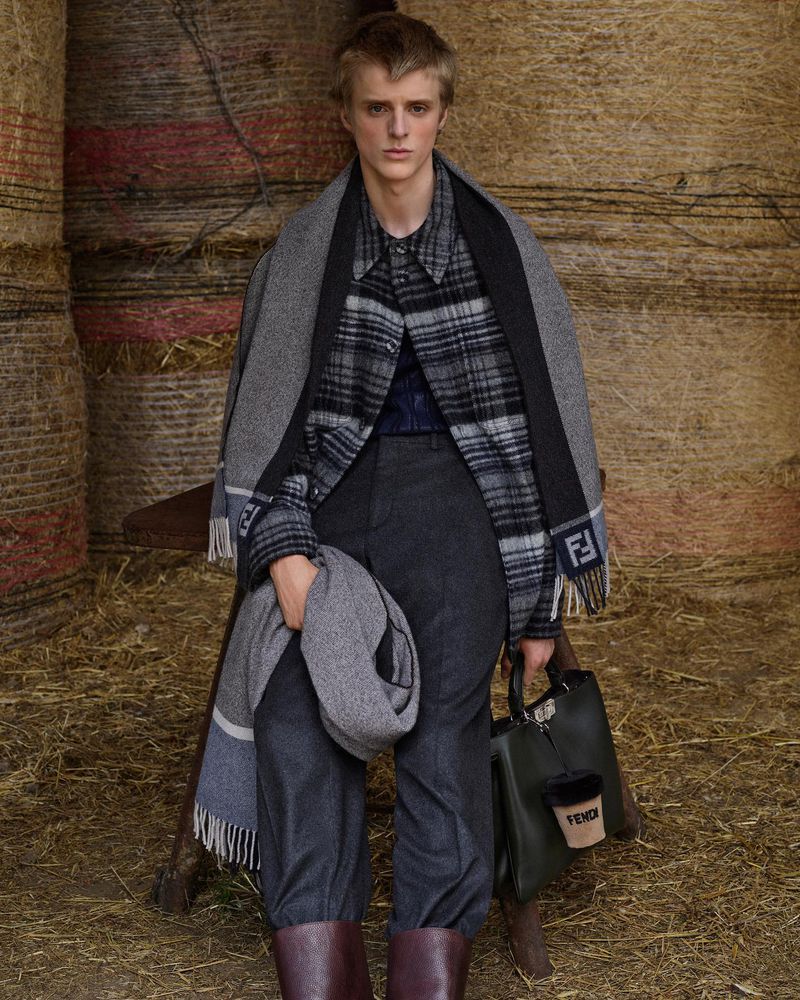 Man wearing a plaid shirt, grey scarf, and boots stands in a hay storage, holding a purse and another scarf; fashion advertisement.
