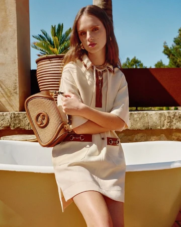 A young, fair-skinned woman wearing a beige linen mini dress poses with a woven handbag, outdoors with potted plants and a vintage bathtub.