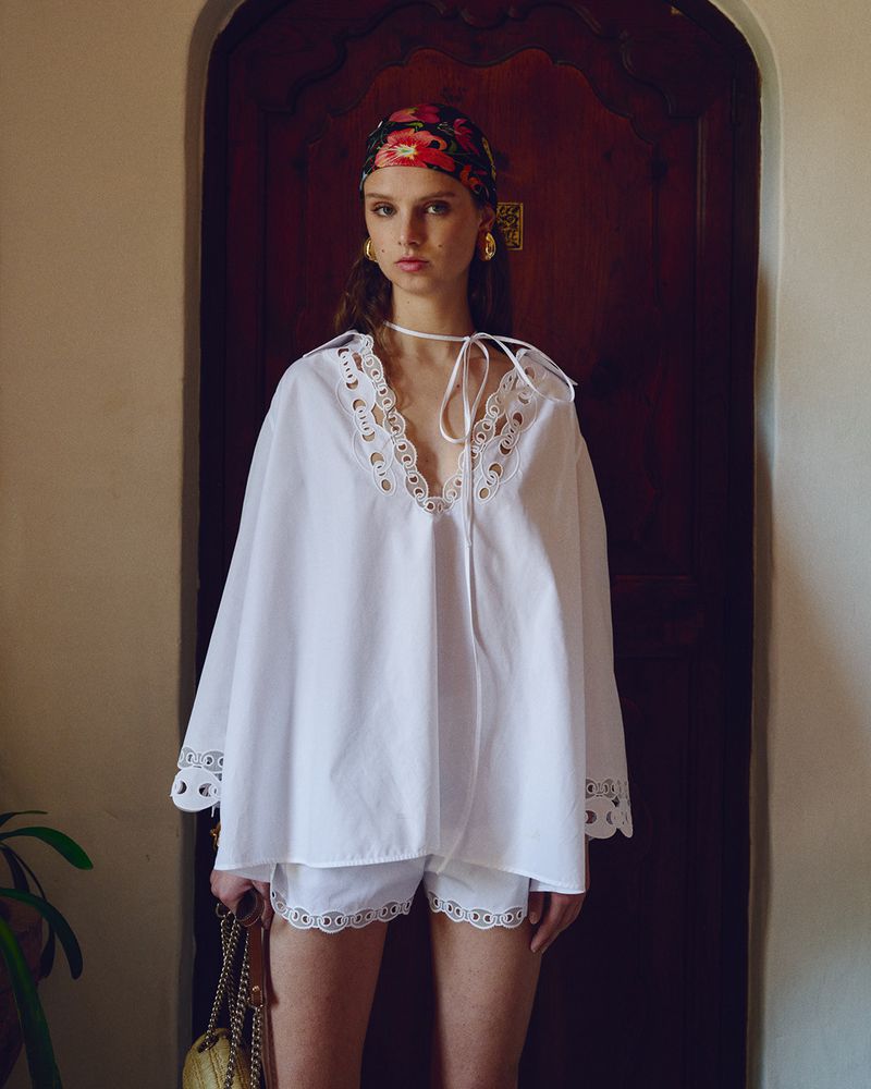 Woman in white tunic top and shorts with floral headscarf stands indoors by dark wood door, accessorized with gold earrings and straw bag.