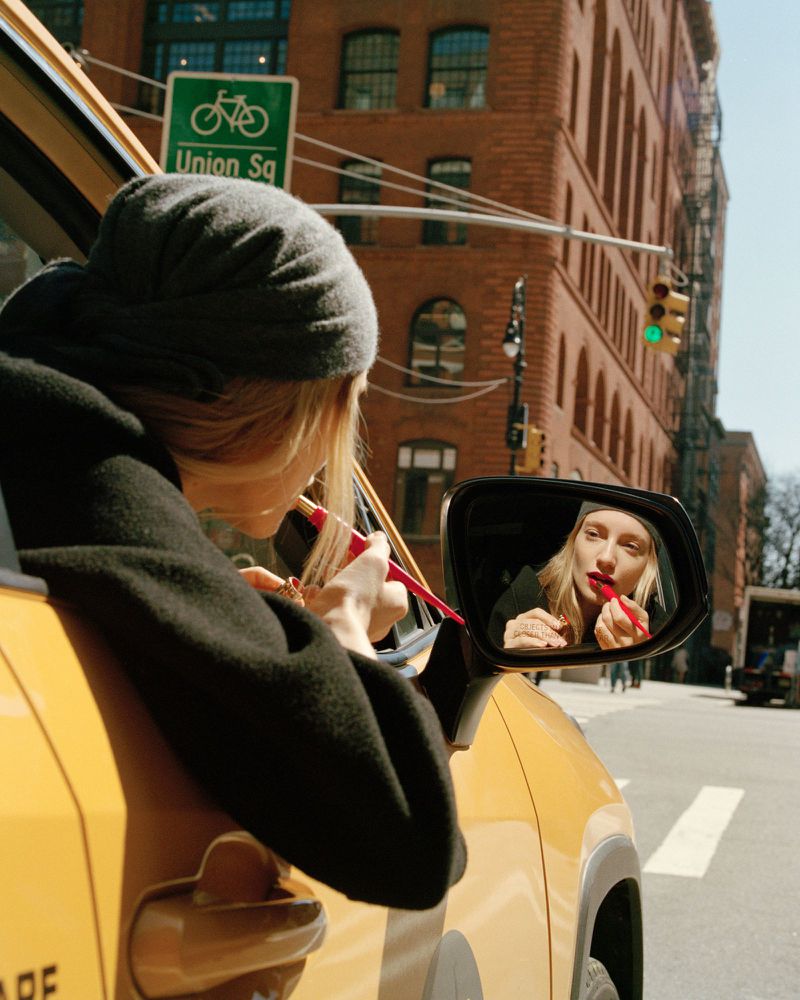 Woman in a yellow car applies red lipstick using the car's side mirror on a city street with brick buildings and a 'Union Sq' sign.