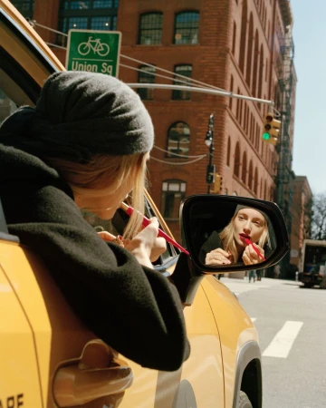 Woman in a yellow car applies red lipstick using the car's side mirror on a city street with brick buildings and a 'Union Sq' sign.