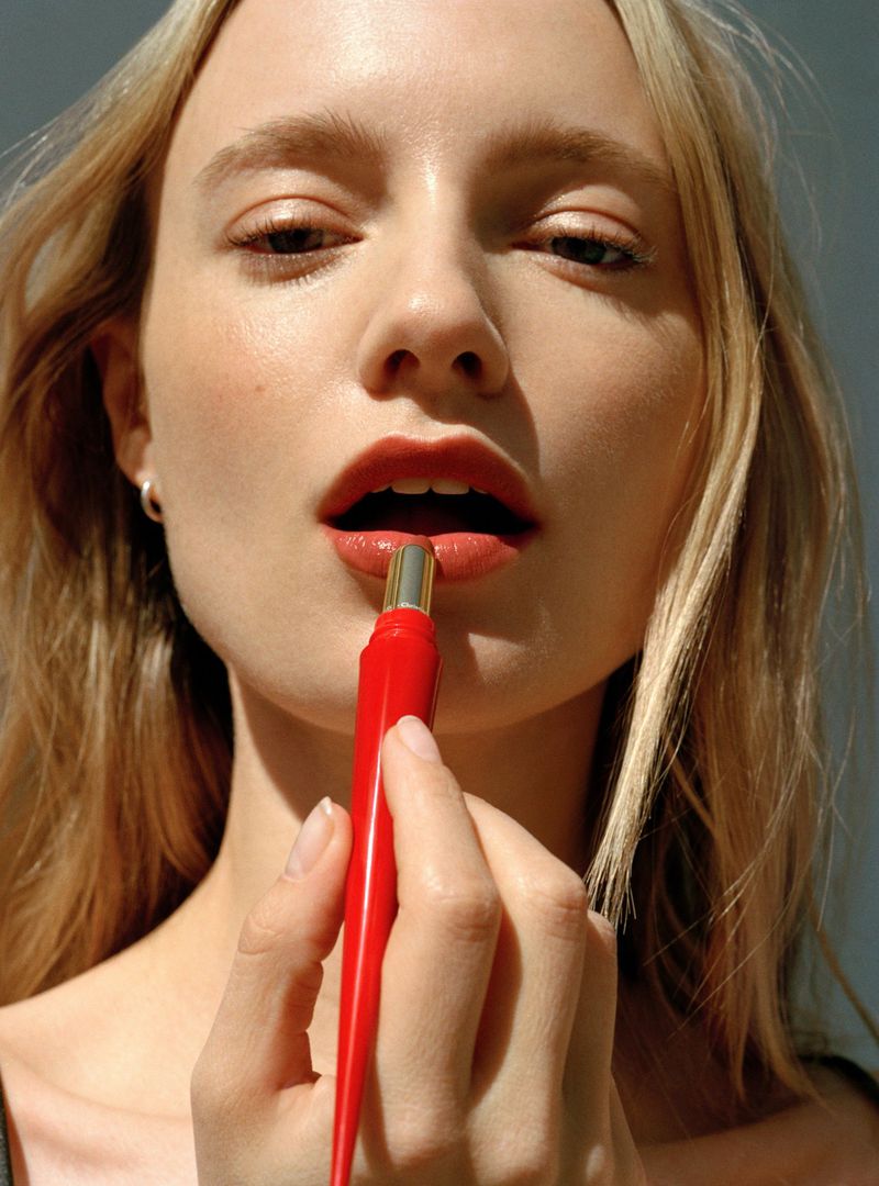 Close-up of a blond woman applying red lipstick with a pointed applicator, showcasing beauty and makeup, with a soft-focus background.