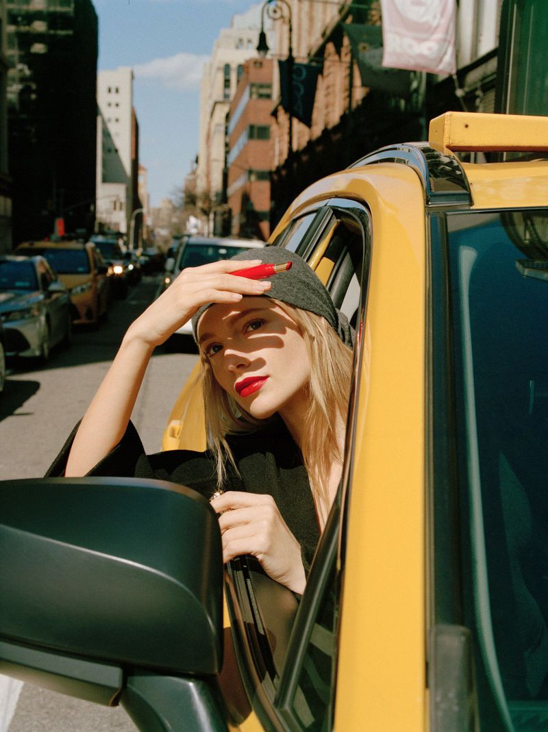 A woman in a yellow taxi cab in a city street wearing a baseball cap, with other city traffic and buildings in the background.